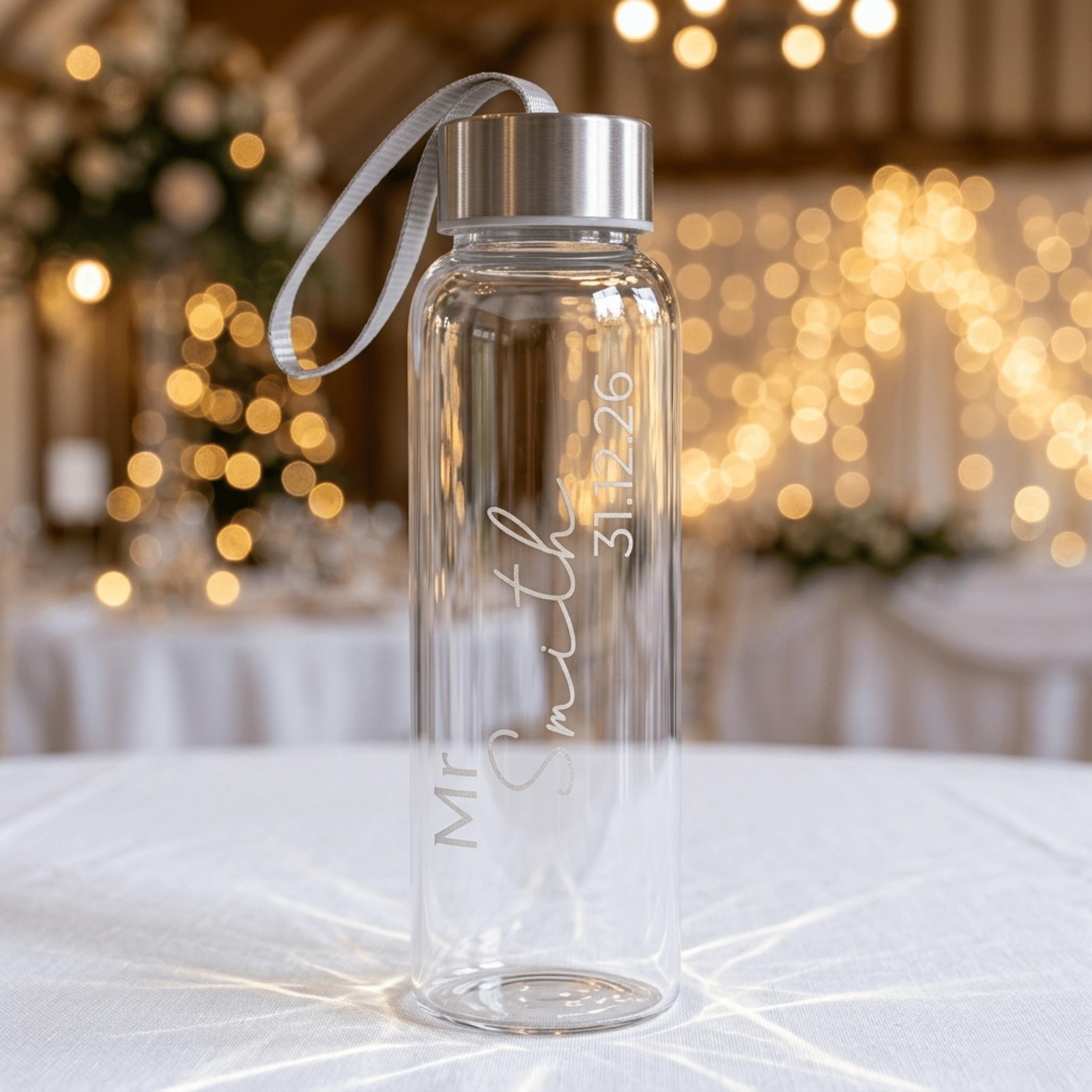 Custom engraved glass water bottle for a groom, featuring "Mr Smith" and a wedding date in elegant vertical script. This personalised gift idea is shown in a festive wedding venue setting with fairy lights in the background.
