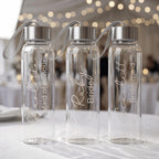 Three personalised glass water bottles for a bridal party featuring "Charlotte Maid of honour", "Ruby Bride", and "Scarlett Bridesmaid" in elegant white script, displayed at a wedding reception.