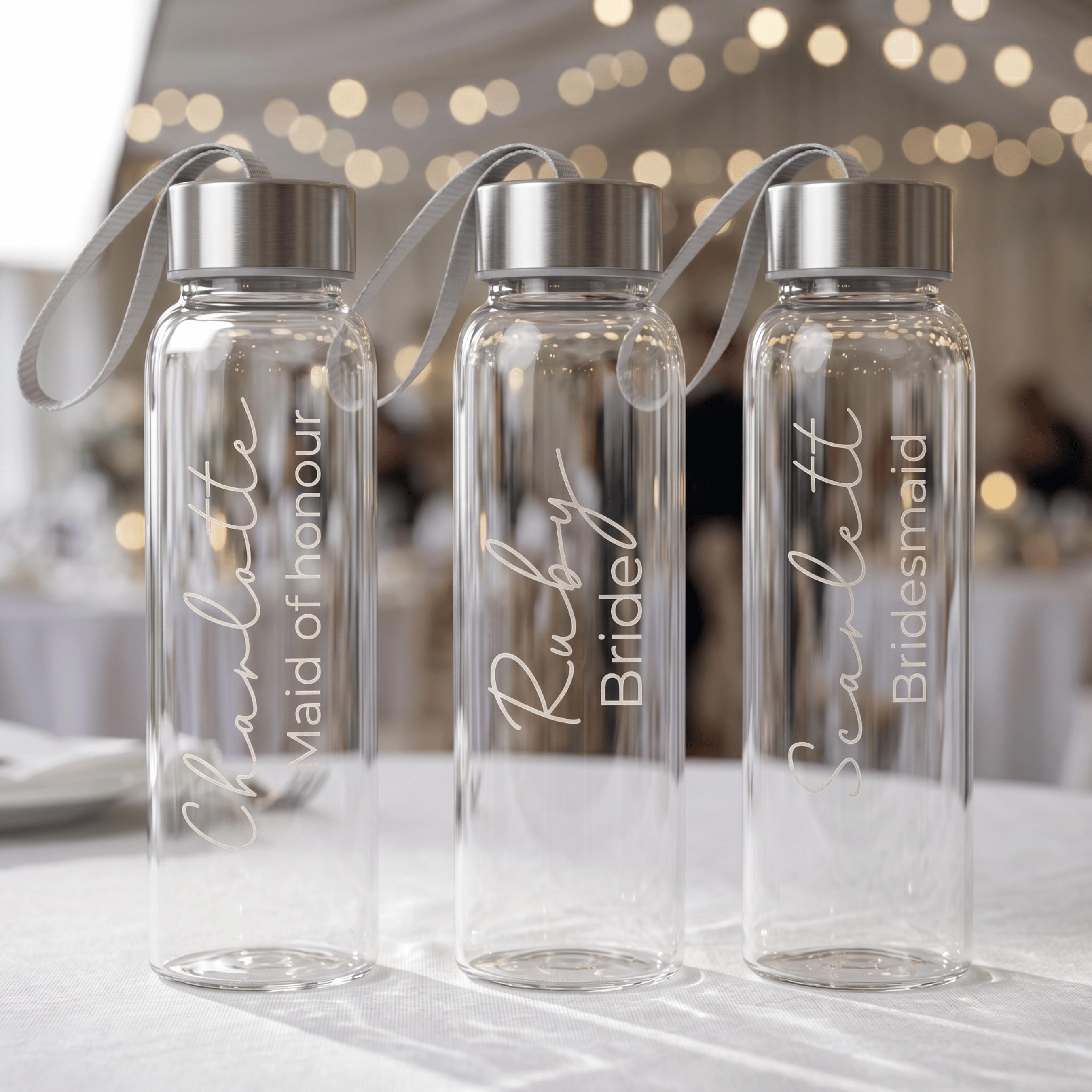 Three personalised glass water bottles for a bridal party featuring "Charlotte Maid of honour", "Ruby Bride", and "Scarlett Bridesmaid" in elegant white script, displayed at a wedding reception.