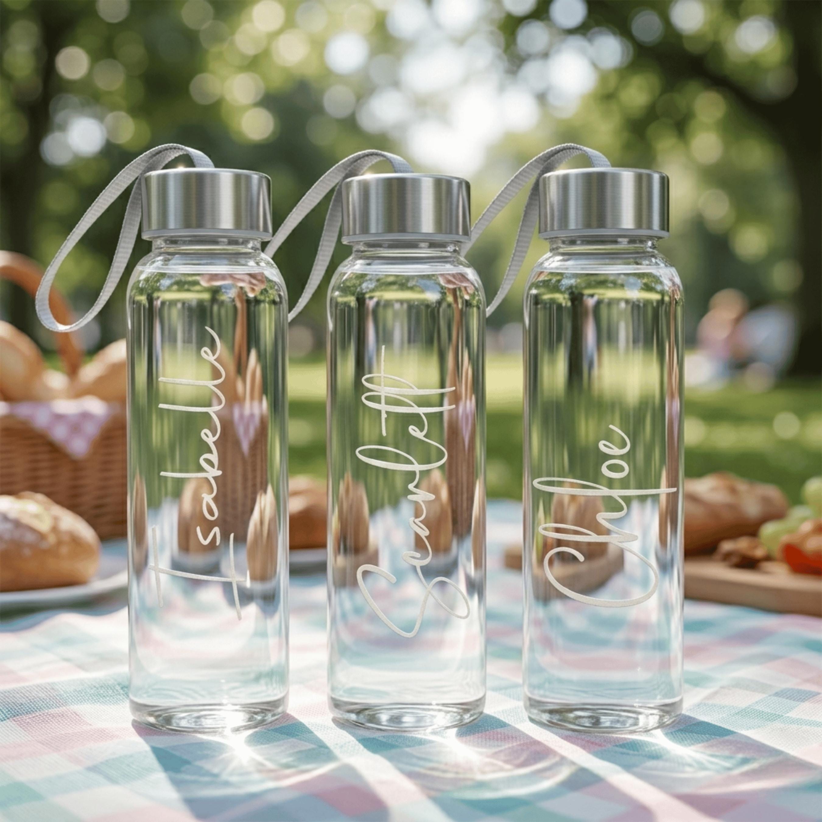 Three personalised glass water bottles with names Isabelle, Scarlett, and Chloe in white script, arranged on a checkered picnic rug in a sunlit outdoor park.