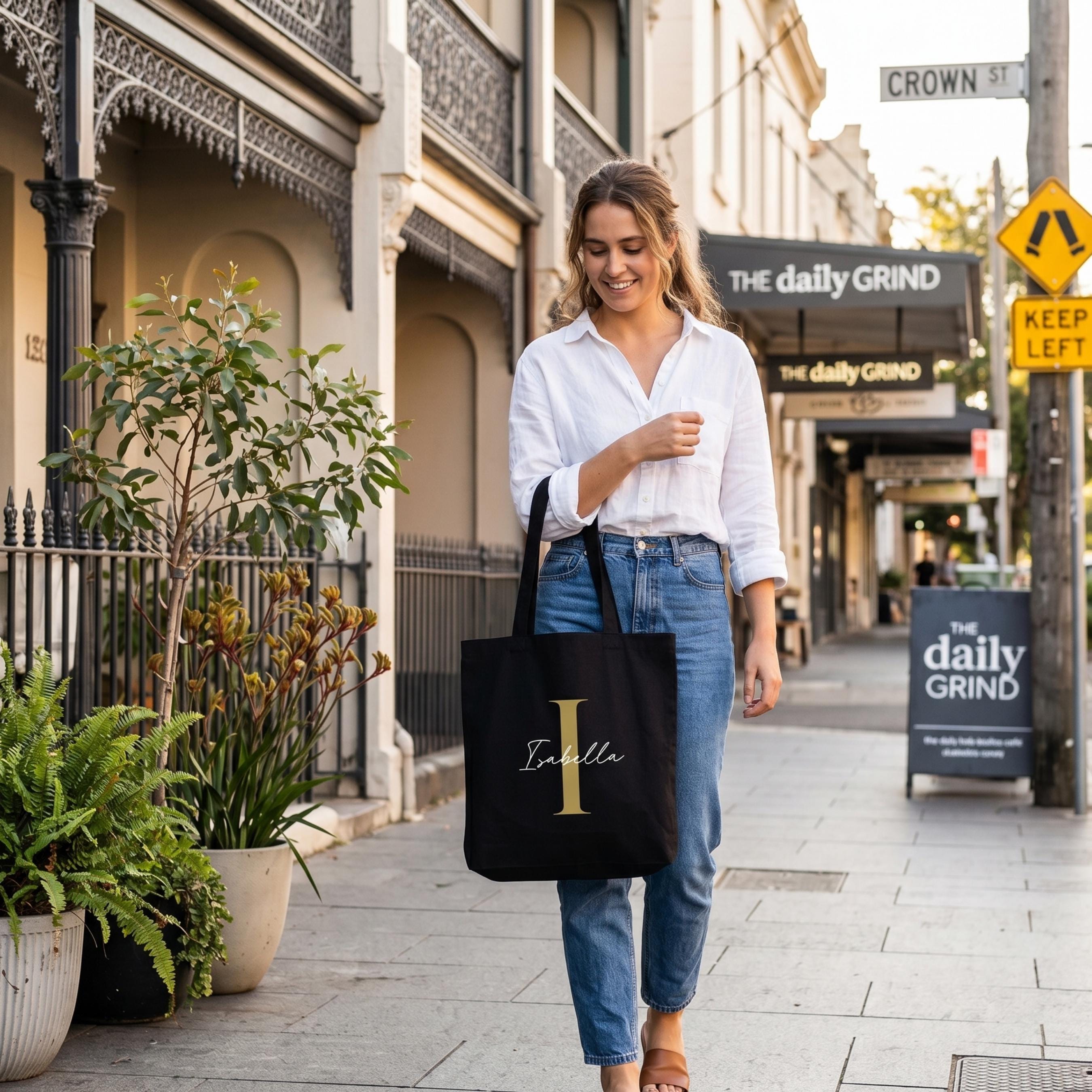Lifestyle shot showing a woman on Crown Street with a black personalised tote bag. This custom shopping bag is detailed with a large gold letter 'I' and a name, perfect as a unique personalised gift for an eco-conscious Australian lifestyle.