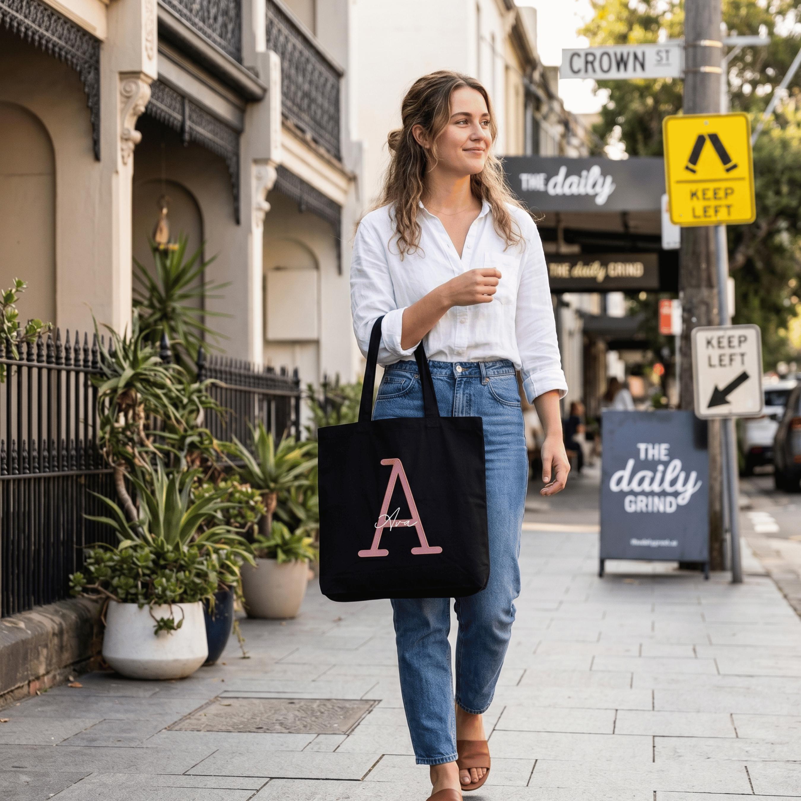 Lifestyle shot showing a woman on Crown Street with a black personalised tote bag. This custom shopping bag is detailed with a large rose gold letter 'A' and a name, perfect as a unique personalised gift for an eco-conscious Australian lifestyle.