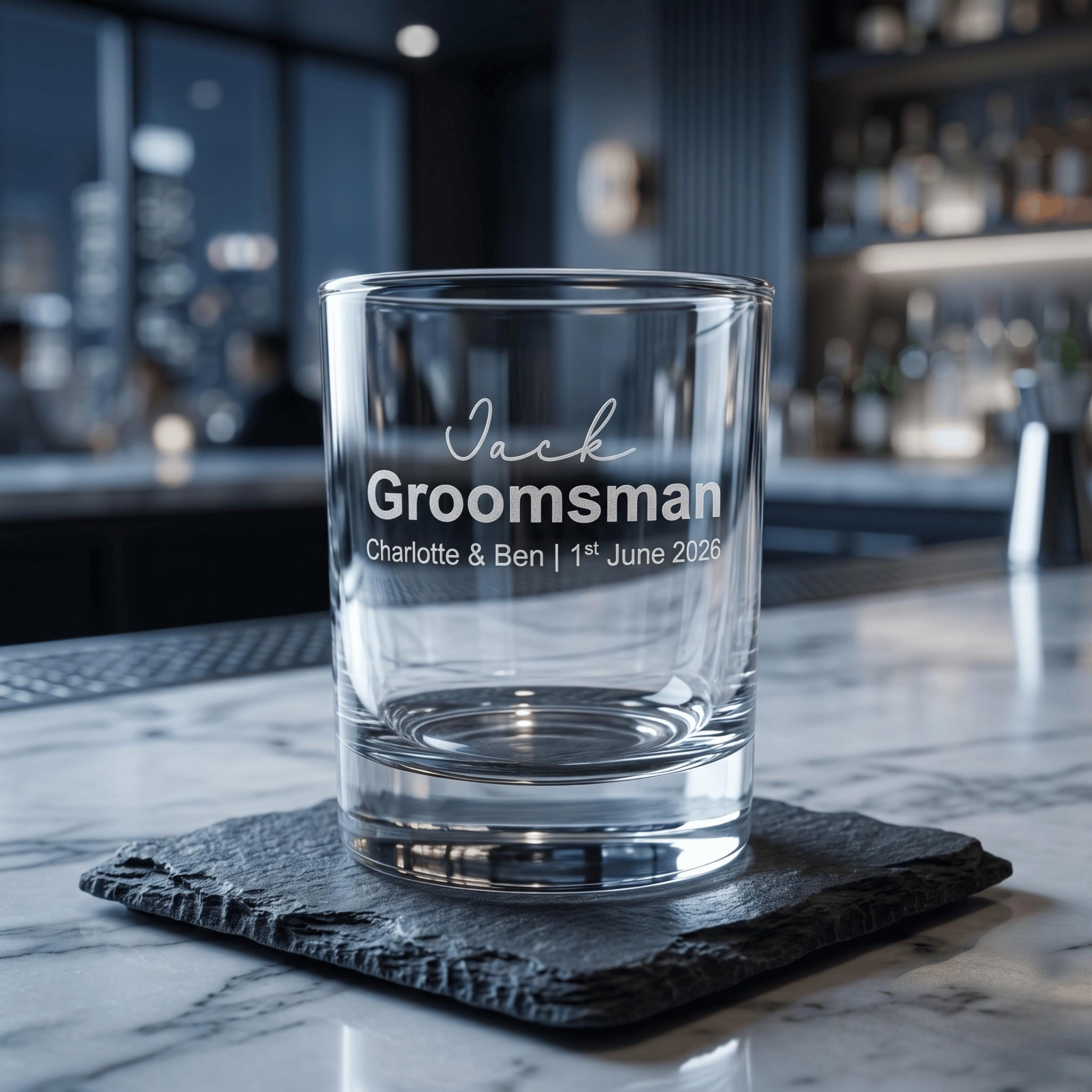 Close-up of a personalised groomsman shot glass engraved with the name "Jack" and wedding details, resting on a rustic slate coaster in a modern bar setting.