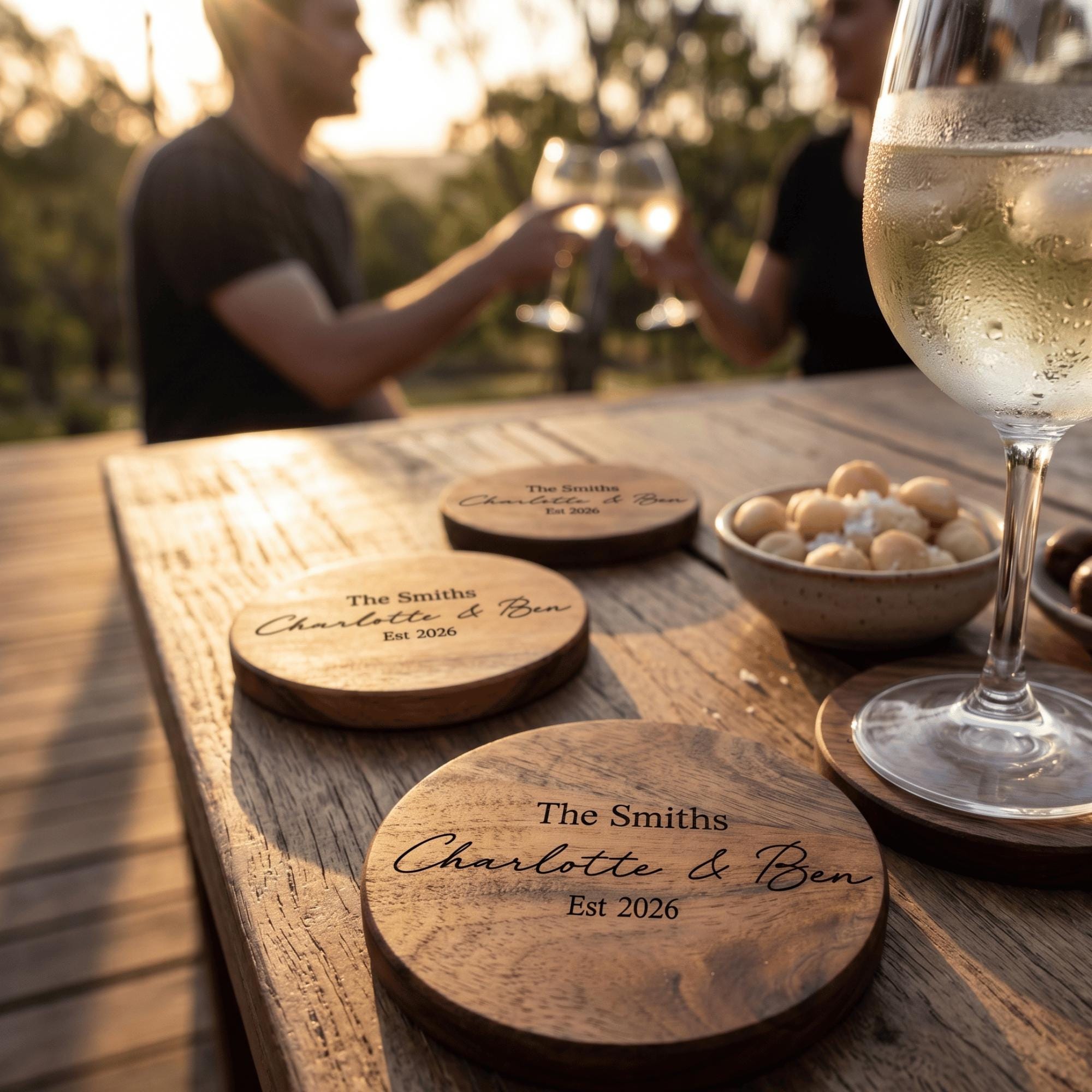 Wooden coasters with engraved names on a wooden table with wine glasses and snacks.