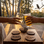 Two people toasting with glasses of beer on a wooden table outdoors during sunset.
