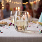 Stemless wine glass with '30 Ava' engraving on a festive table with blurred people in the background.