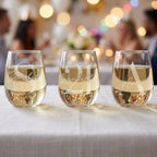 Three stemless wine glasses with names engraved on them, filled with a white beverage, on a table with a blurred festive background.