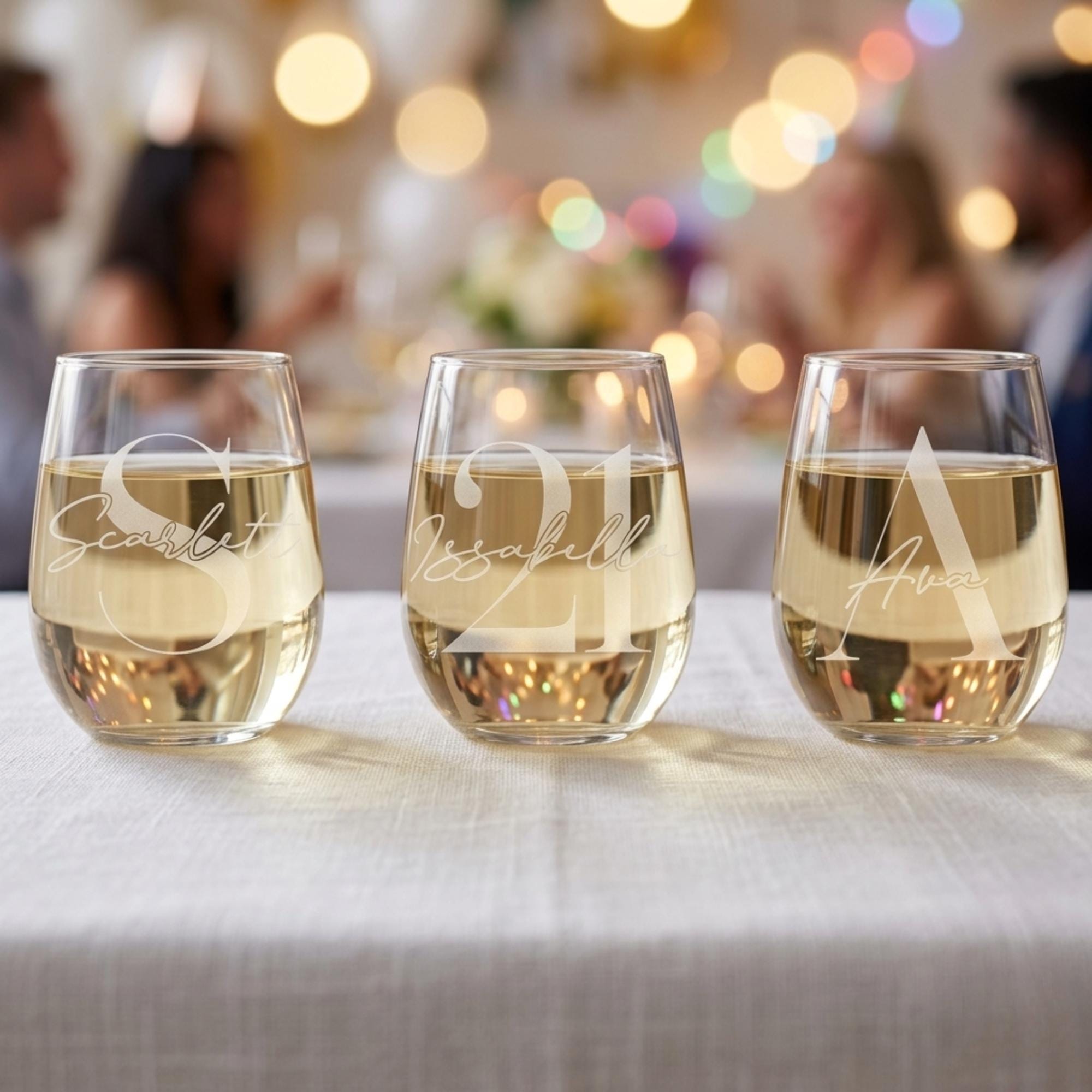Three stemless wine glasses with names engraved on them, filled with a white beverage, on a table with a blurred festive background.