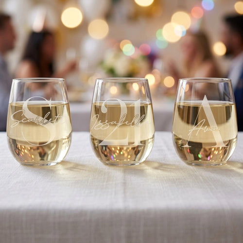 Three stemless wine glasses with names engraved on them, filled with a white beverage, on a table with a blurred festive background.