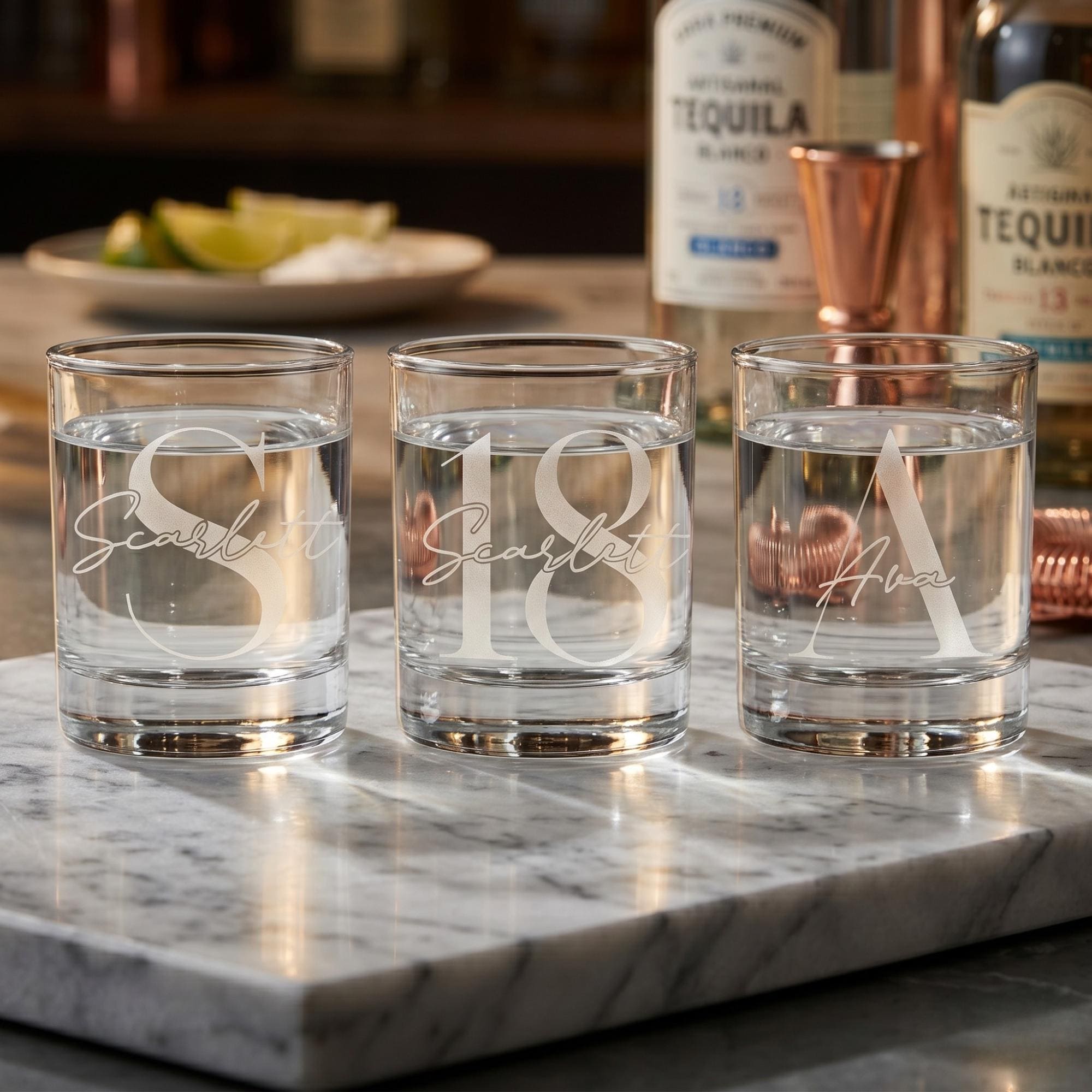 Three personalized shot glasses on a marble surface with tequila bottles in the background.