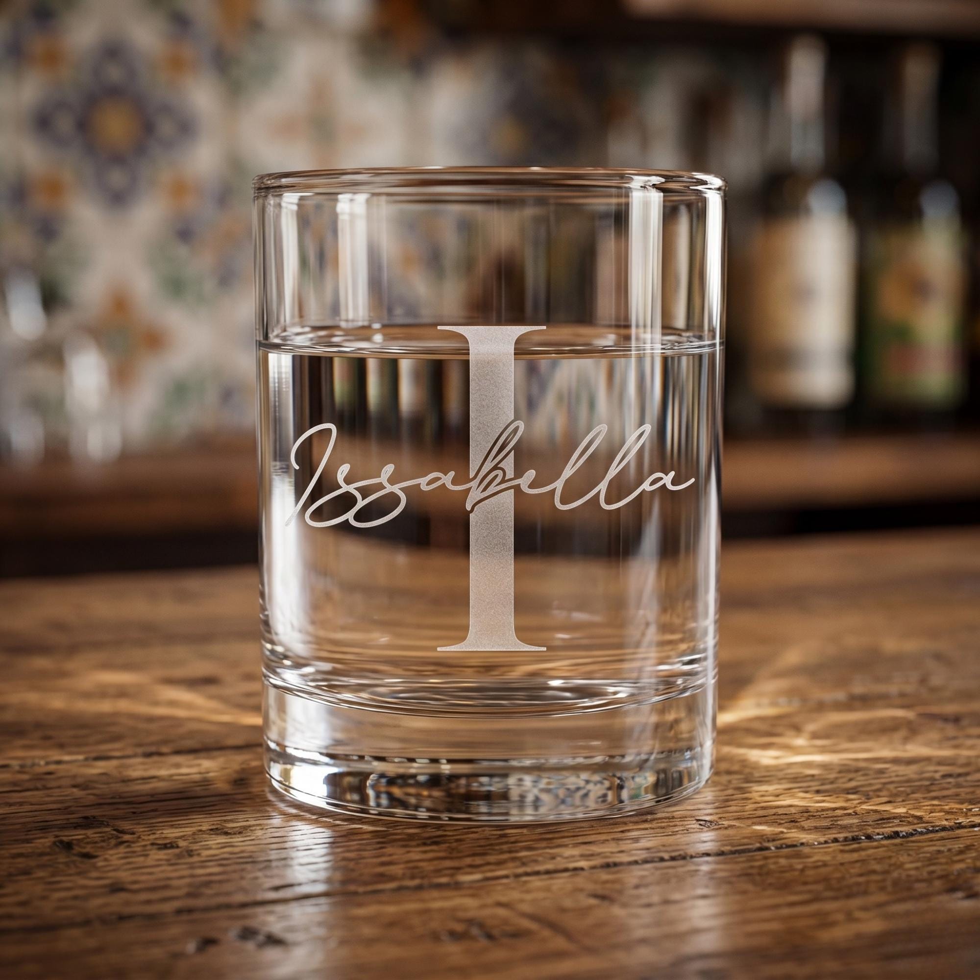 Clear glass with 'Issabella' engraving on a wooden surface with blurred bottles in the background