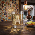 Glass of alcohol with 'Ava' engraving on a wooden table with decorative tiles and lighting in the background.
