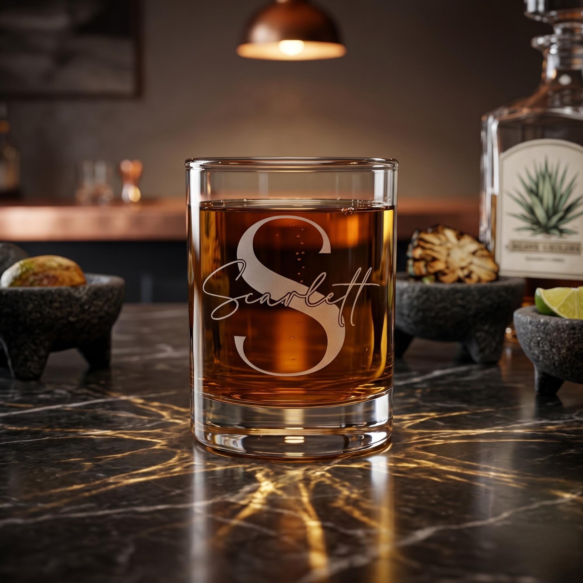 Glass of amber liquid with 'Scarlett' engraving on a dark marble surface, with a bottle and ingredients in the background.