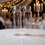 A pair of personalised glass water bottles engraved with "Mr Smith" and "Mrs Smith" and their wedding date 31.12.26. These custom-made drink bottles are displayed on a white tablecloth at a wedding reception with elegant warm bokeh lighting.