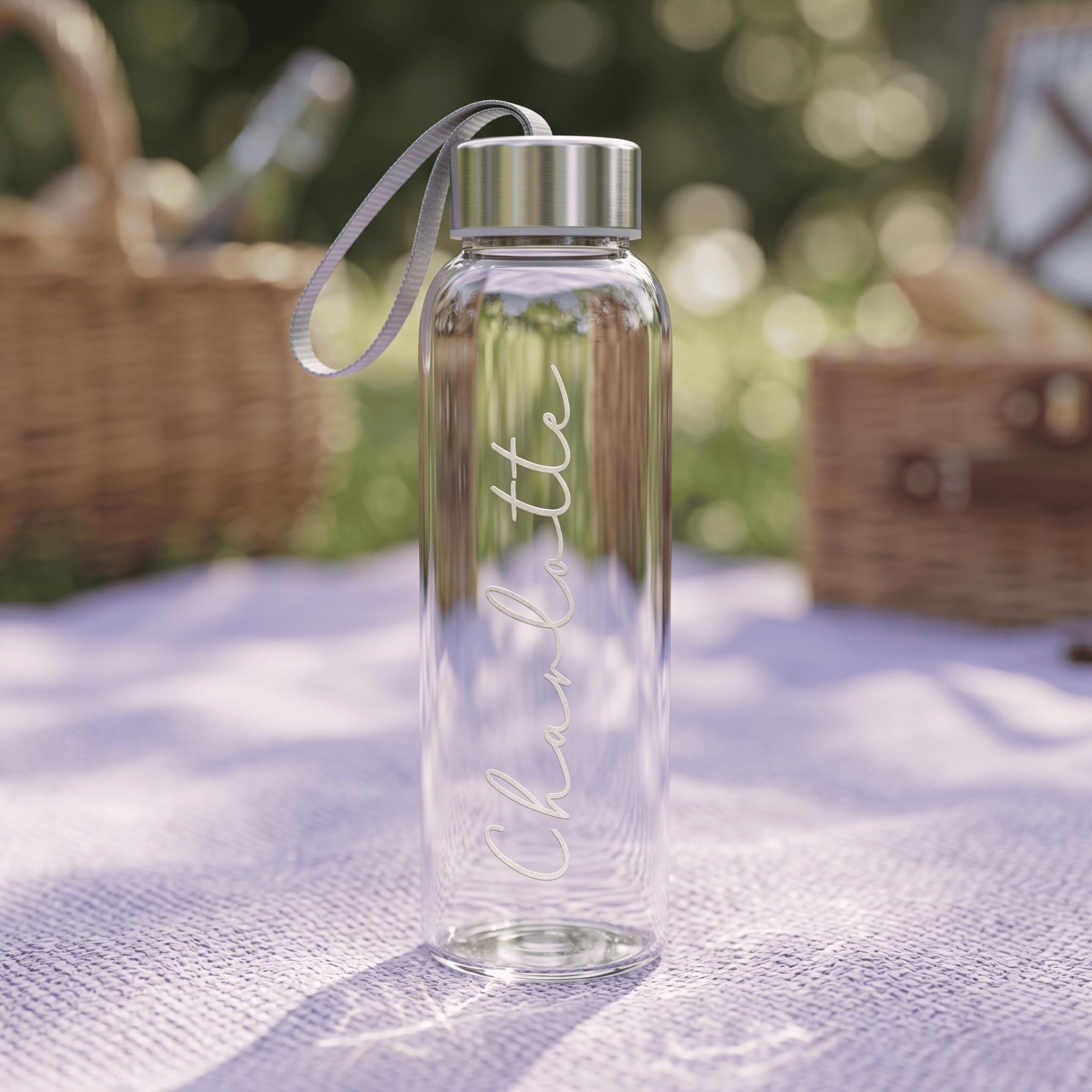 A custom-made glass drink bottle featuring the name "Charlotte" in elegant cursive, resting on a purple picnic blanket with a wicker basket in the background.