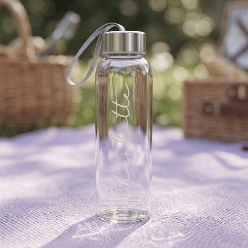 A custom-made glass drink bottle featuring the name "Charlotte" in elegant cursive, resting on a purple picnic blanket with a wicker basket in the background.