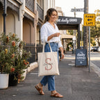 A woman strolling down a city pavement , carrying a cream-coloured personalised canvas tote bag. The bag is customised with a large tan letter 'S' and the name 'Scarlett', representing a high-quality, personalised Australian accessory.