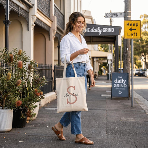 A woman strolling down a city pavement , carrying a cream-coloured personalised canvas tote bag. The bag is customised with a large tan letter 'S' and the name 'Scarlett', representing a high-quality, personalised Australian accessory.