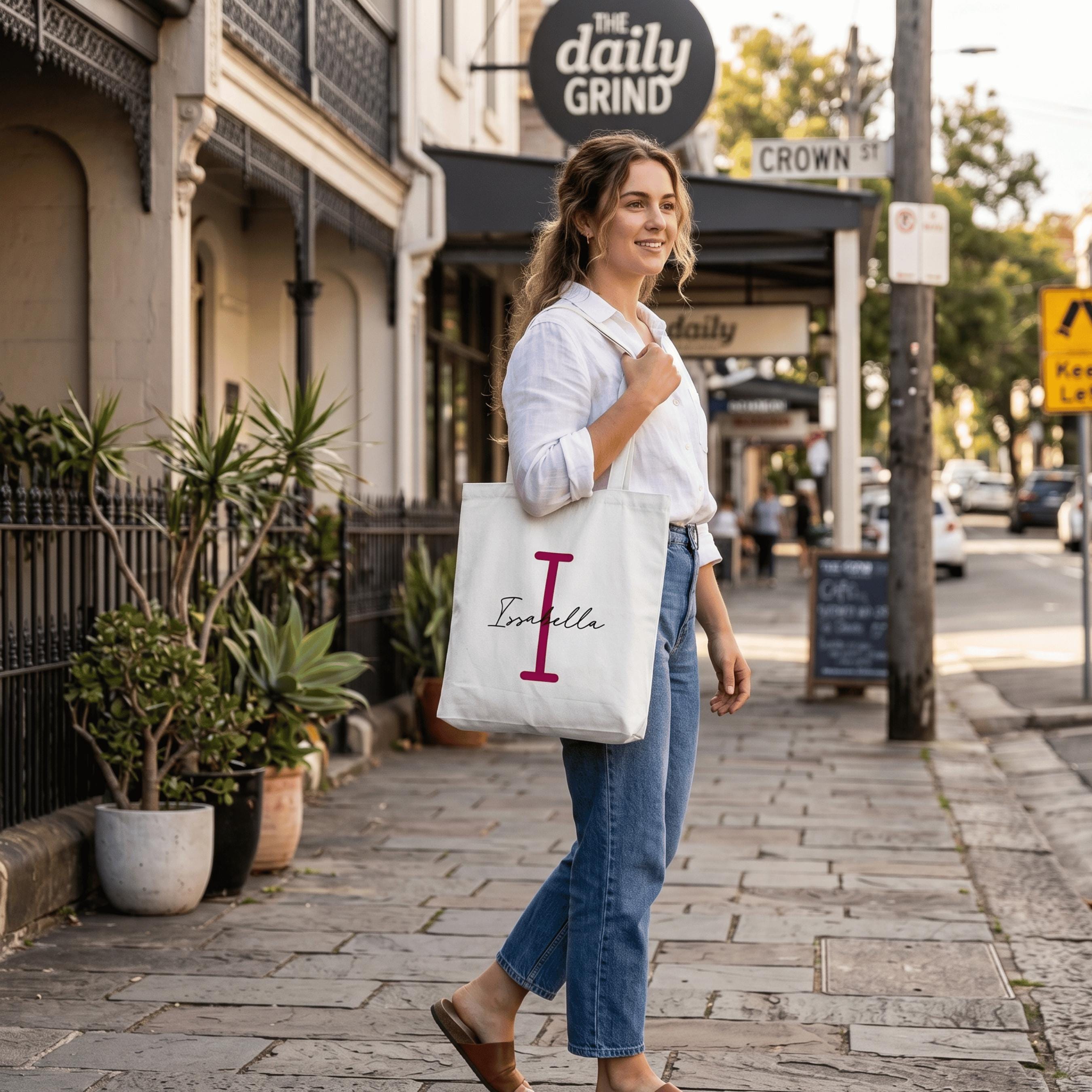 A woman walking past a cafe in a sunlit Australian street, carrying a white-coloured personalised tote bag . The custom canvas bag features a large pink initial 'I' and the name 'Isabelle' in elegant script, showcasing a stylish and sustainable fashion choice.