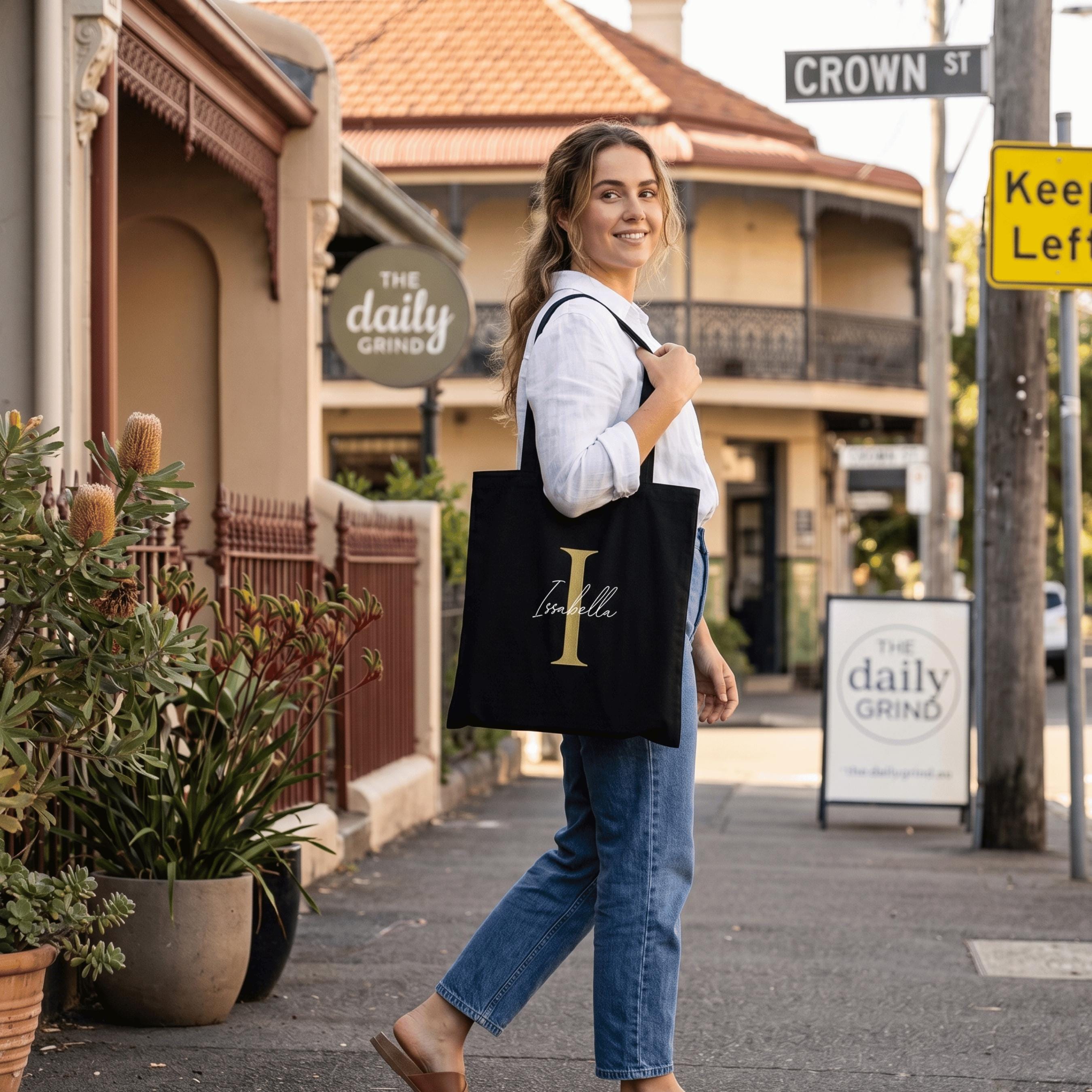Lifestyle shot showing a woman on Crown Street with a black personalised tote bag. This custom shopping bag is detailed with a large pink letter 'I' and a name, perfect as a unique personalised gift for an eco-conscious Australian lifestyle.