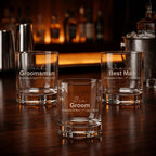 A set of three personalised whisky glasses for a wedding, engraved with "Jack Groomsman", "Ben Groom", and "James Best Man" alongside a wedding date. The glasses are displayed on a dark timber bar with warm, ambient lighting.