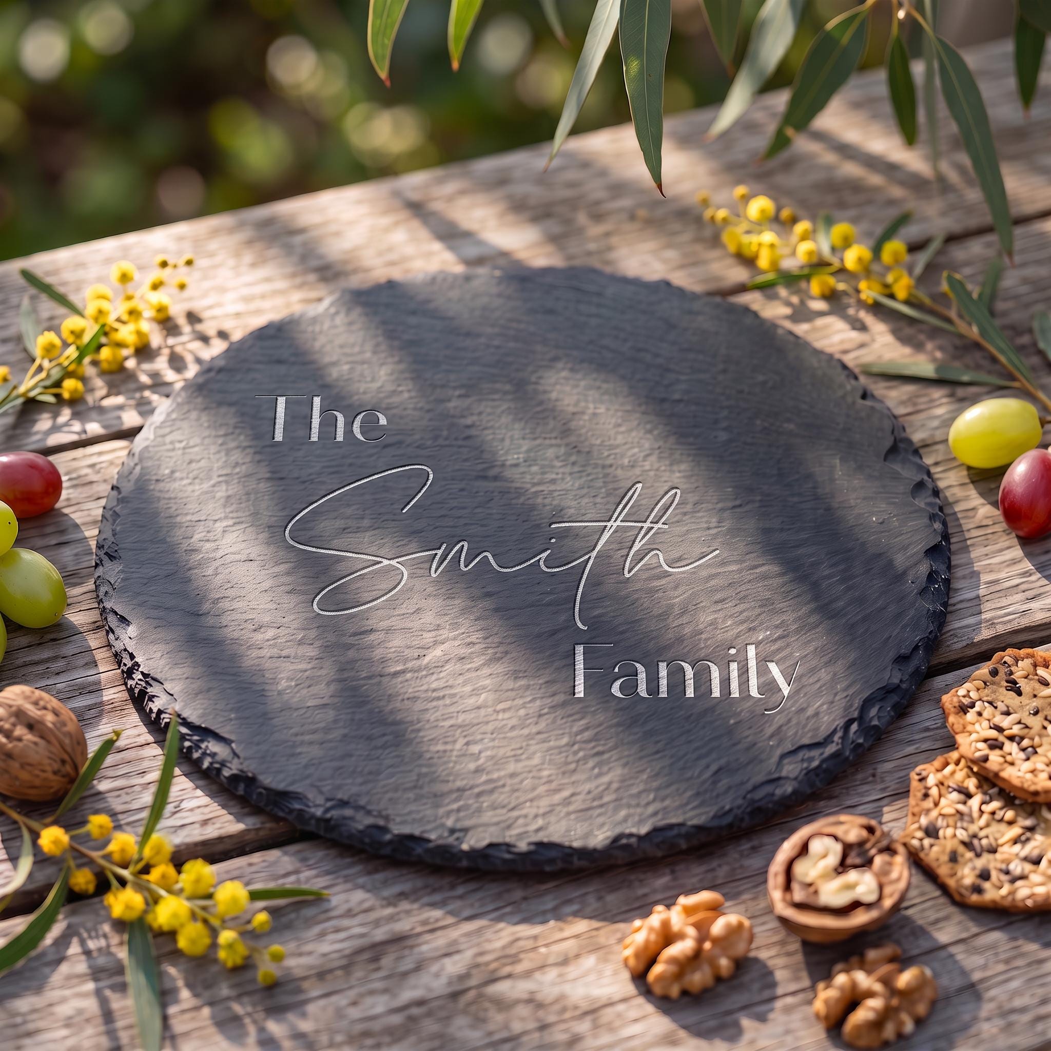 Personalised round slate cheese board engraved with The Smith Family in modern sans and calligraphy script on rustic timber table.