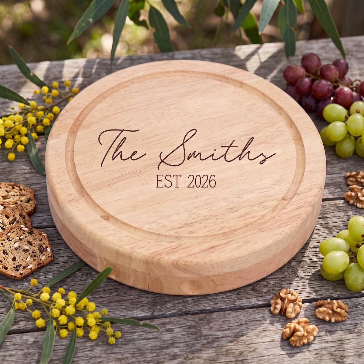 Personalised laser engraved wooden cheese board with juice groove reading The Smiths EST 2026 in delicate script on an outdoor timber table with fresh fruit and crackers.