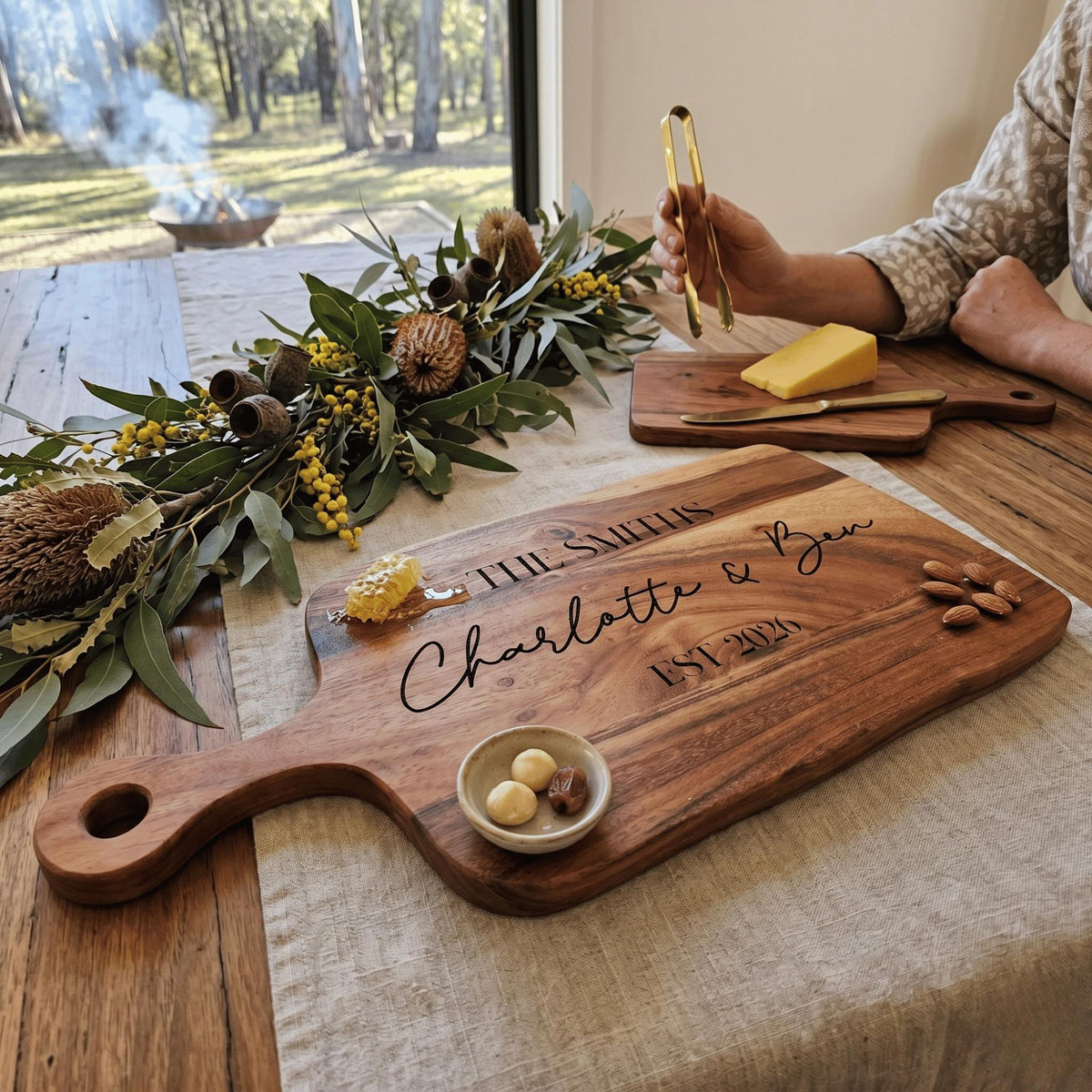 Long acacia wood charcuterie grazing paddle board with handle personalised with The Smiths Charlotte and Ben EST 2026 with cheese and native leaves.