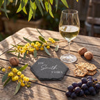 Personalised hexagon slate drinks coaster engraved with THE Smith FAMILY in all caps serif and delicate script beside a wine glass on timber table.
