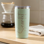A mint green insulated travel tumbler with a clear lid and the word 'Scarlet' in metallic silver engraving, standing on a wooden countertop with a blurred coffee maker and folded cloth in the background.