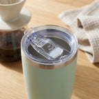 A mint green insulated tumbler with a clear lid and clear straw insert, shown on a wooden tabletop with a coffee maker and cloth in the background.