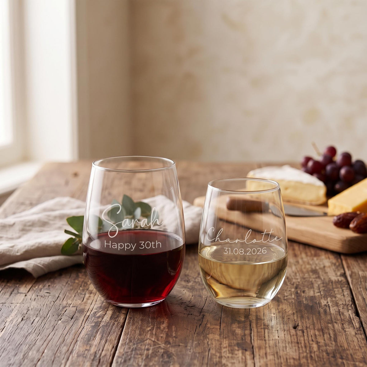 Personalised engraved stemless wine glass displaying custom names and milestone dates on a rustic wooden table with cheese and grapes.