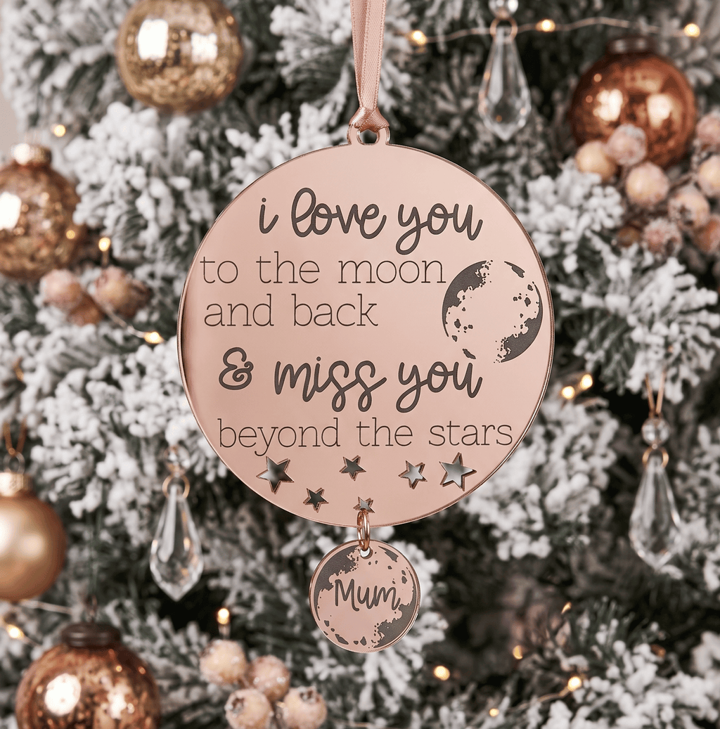 A round rose gold Christmas ornament hanging on a decorated Christmas tree. The ornament features engraved text that reads 'i love you to the moon and back & miss you beyond the stars' along with moon and star illustrations, and a smaller dangling charm engraved with 'Mum'.