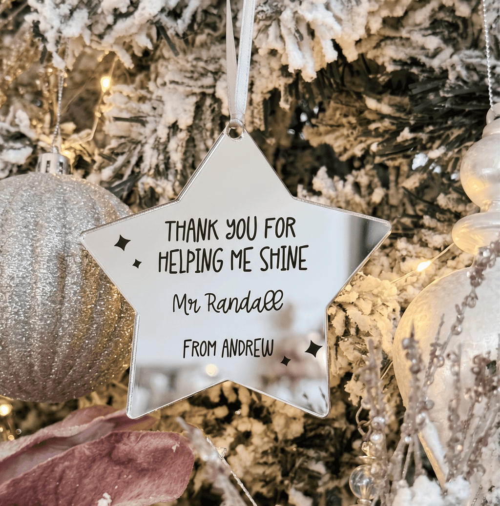 A personalised star-shaped Christmas ornament hanging on a decorated tree. The ornament is silver with engraved text that reads, 'THANK YOU FOR HELPING ME SHINE Mr.