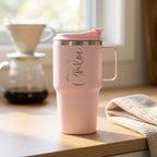 A light pink insulated travel mug with a matching lid and handle sits on a wooden table in a sunlit kitchen setting. The mug has a silver metallic script name 'Chloe' printed on the front.