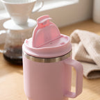 A light pink insulated mug with a matching pink lid and flip-up straw cover, featuring a stainless steel band near the rim, set on a wooden table with a coffee maker and cloth in the background.