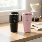 Two insulated travel tumblers with lids and handles sit on a wooden table near a window. One tumbler is matte black with the name 'Will' in silver script, and the other is matte pink with the name 'Chloe' in silver script.